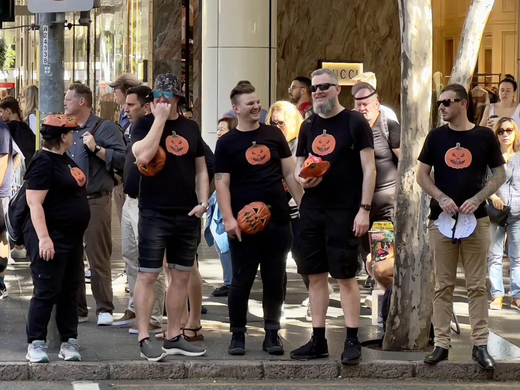 A group of adults in matching black T-shirts printed with jack-o'-lantern faces on a city street in Brisbane