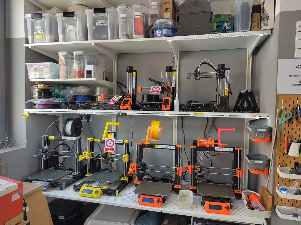 A small 3D printer farm of seven desktop filament printers on shelves at a hackerspace in Wroclaw, Poland