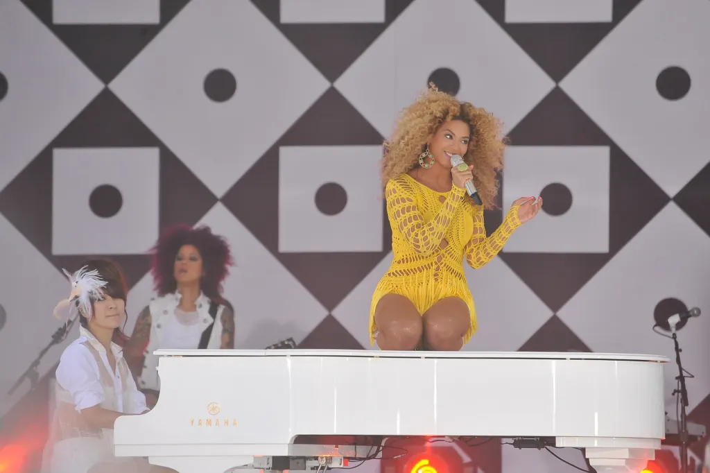 Beyoncé in a yellow outfit sings into a microphone while seated on a white grand piano at an outdoor concert in Central Park, New York, in 2011