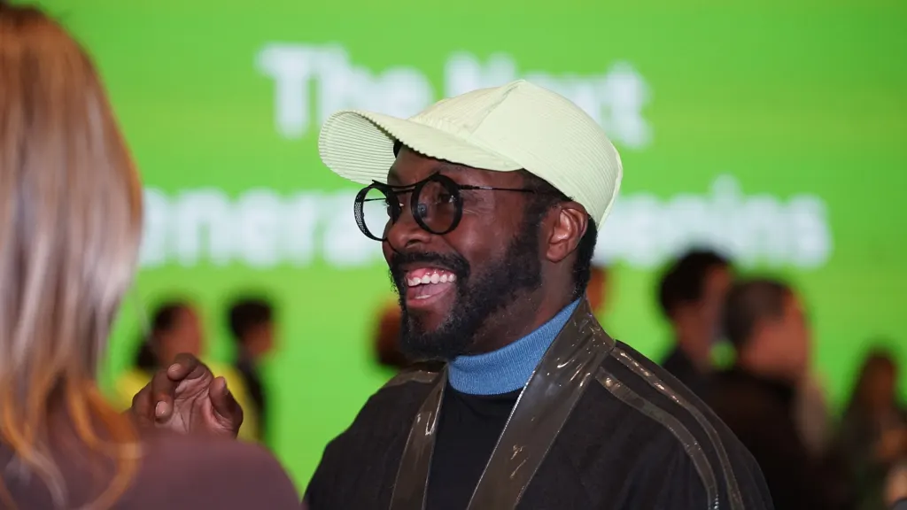 will.i.am, smiling in a pale cap and round glasses, talking to a woman in front of a green screen at CES 2026 in Las Vegas