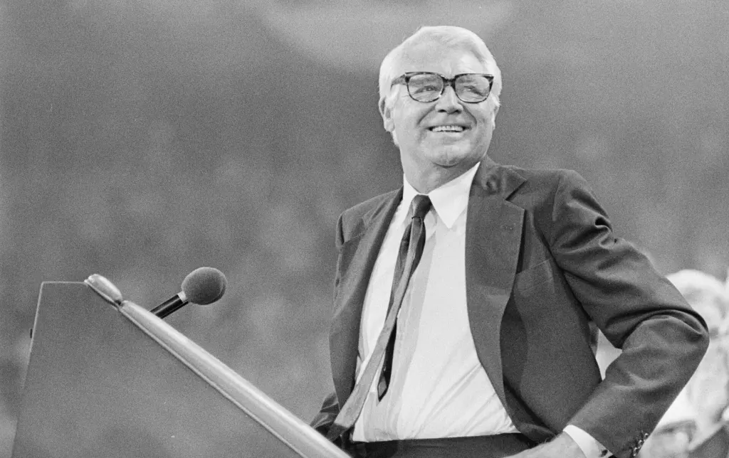 Black-and-white photo of a smiling, white-haired Cary Grant in glasses and a dark suit, standing at a podium at the 1976 Republican National Convention in Kansas City, Missouri
