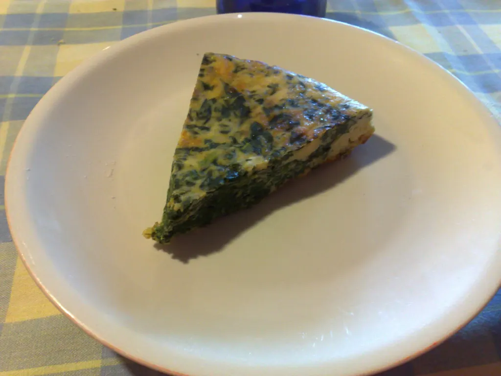 A wedge of Sicilian-style frittata with spinach and onion on a white plate over a blue checked tablecloth