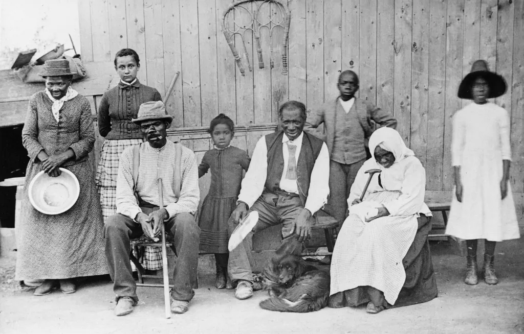 Nineteenth-century group photograph of Harriet Tubman, standing at far left with her hand on a chair, beside family members and neighbors in front of a wooden building
