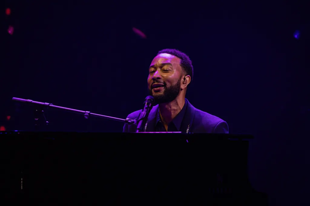 John Legend smiling and singing at a microphone behind a piano under purple stage lights at the Montreux Jazz Festival in July 2026