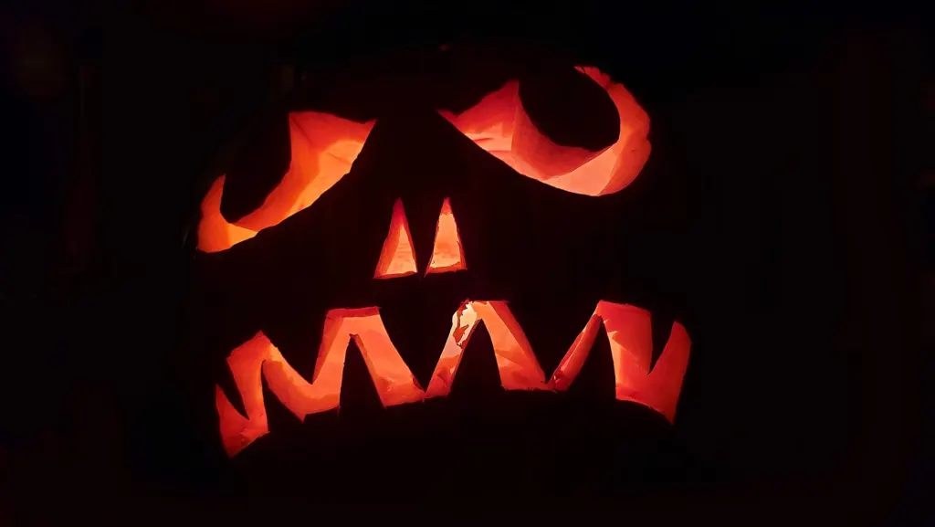 A carved jack-o'-lantern with jagged teeth and slanted eyes glowing orange against a dark porch at night