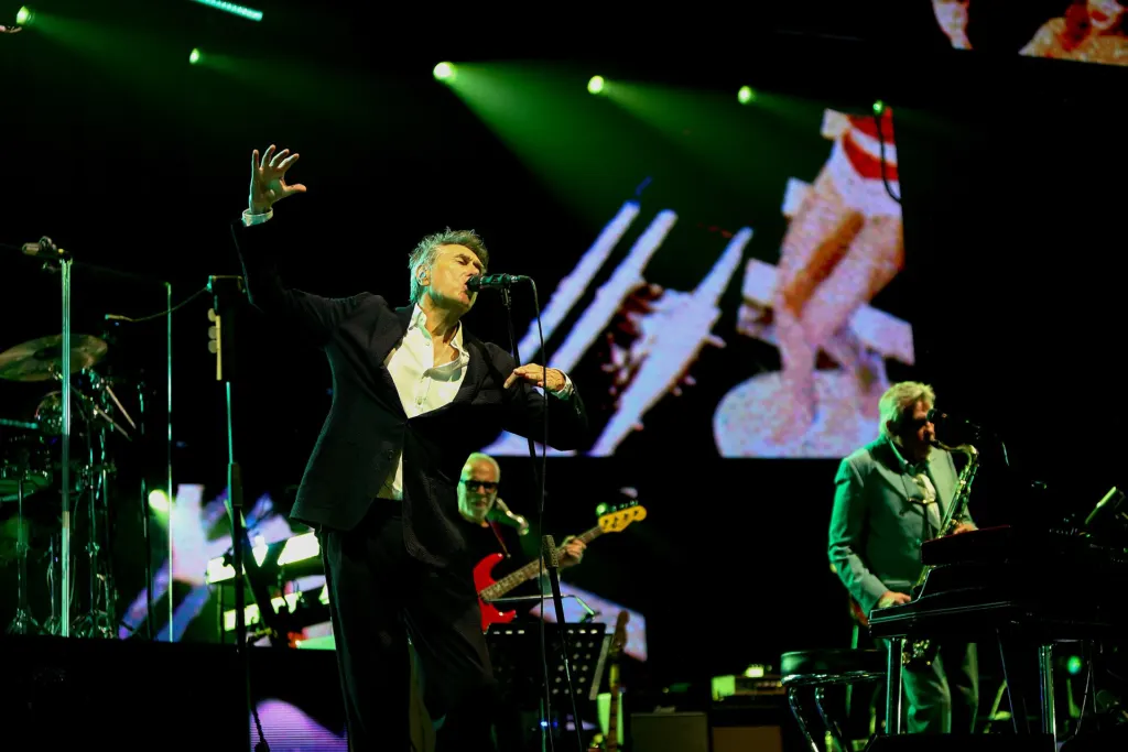Bryan Ferry sings with one arm raised while a saxophonist plays at right during a 2022 Roxy Music concert in Austin, Texas, with green stage lights behind them