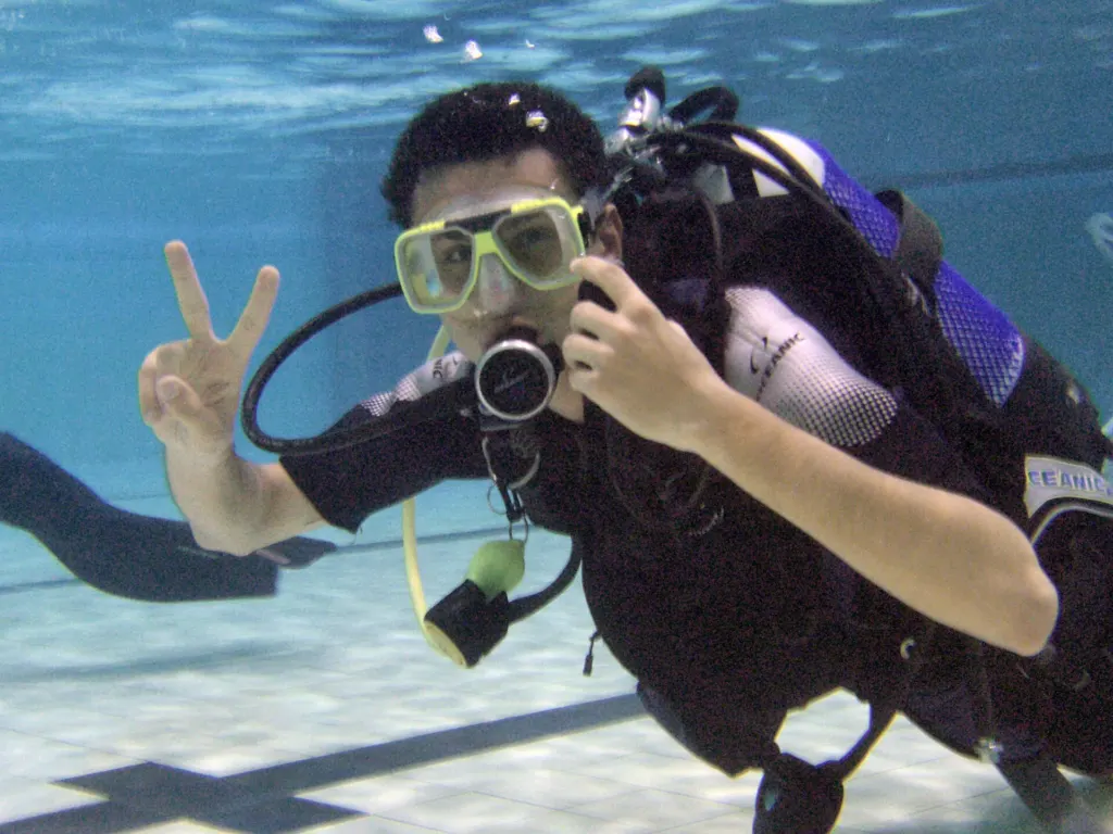 A scuba diver in yellow mask and full gear underwater near the tiled floor of a swimming pool, making a peace sign