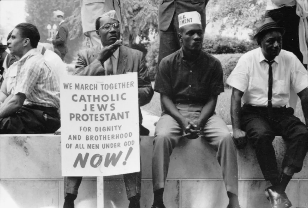 Participants in the 1965 Selma to Montgomery civil rights march rest on a wall; one holds a placard calling for dignity and brotherhood