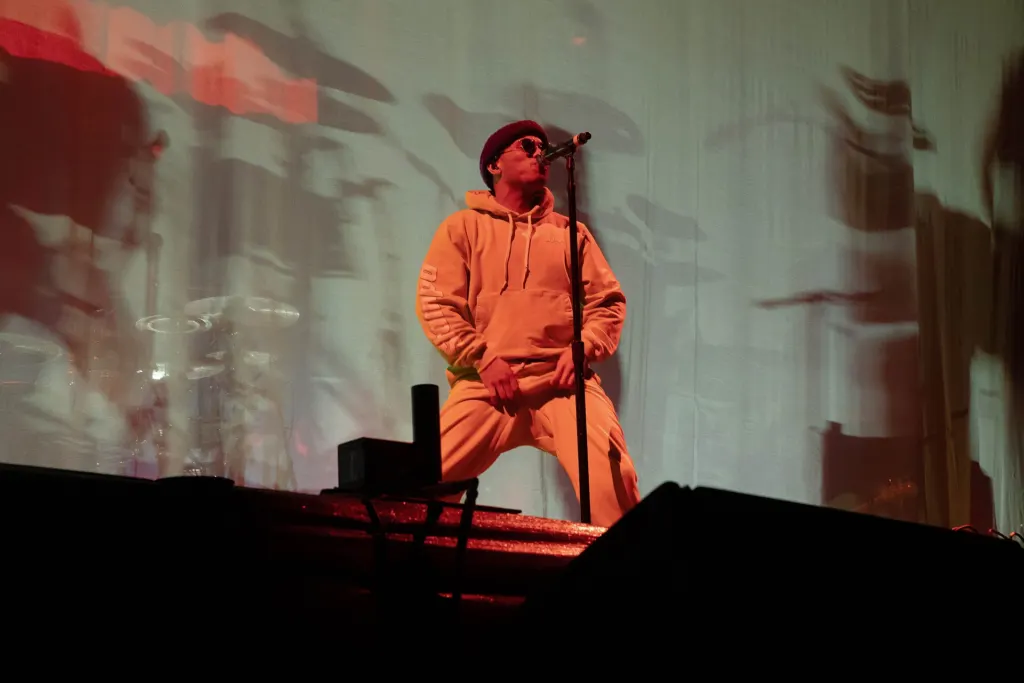 Anderson .Paak in an orange hoodie and dark hat standing at a microphone stand on a concert stage, with a projected image behind him