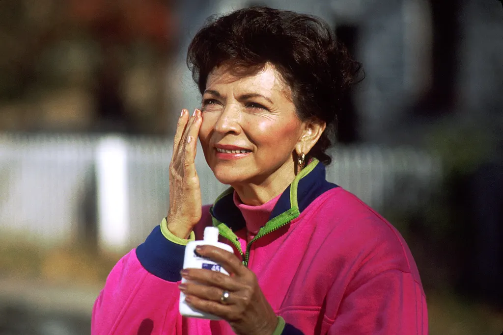 A woman in a hot pink jacket rubs sunscreen onto her face outdoors