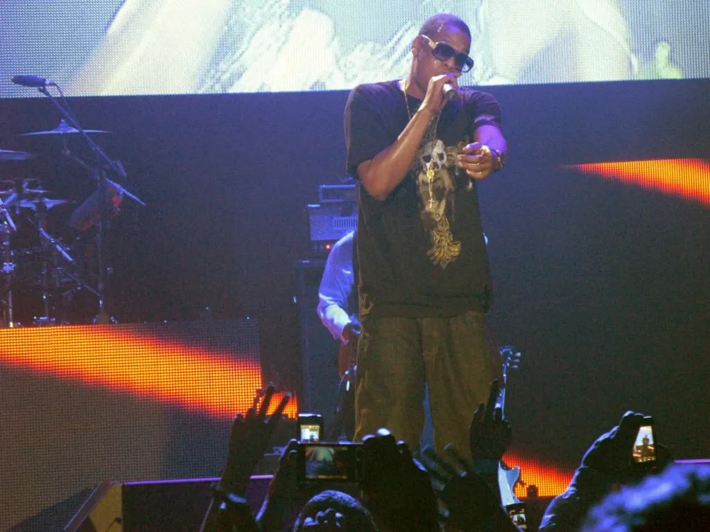 Jay-Z in sunglasses and a black T-shirt performing on a concert stage in 2008 as fans hold up phones in the foreground