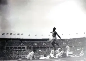 Jesse Owens in midair during the long jump at the Berlin Olympic Stadium in 1936, with officials on the infield and a packed stand and flags behind him