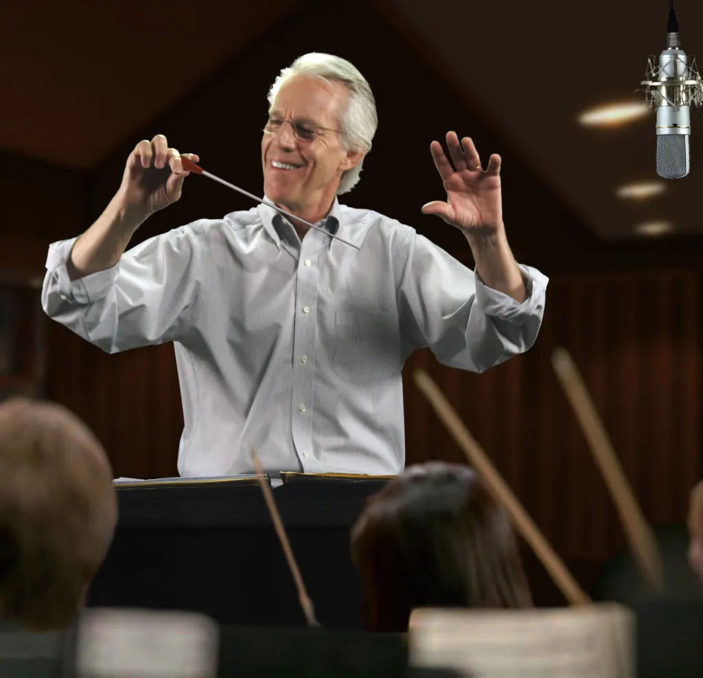 Jim Ed Norman, gray-haired and wearing a light blue shirt, conducting with a baton in a recording studio beside a hanging microphone