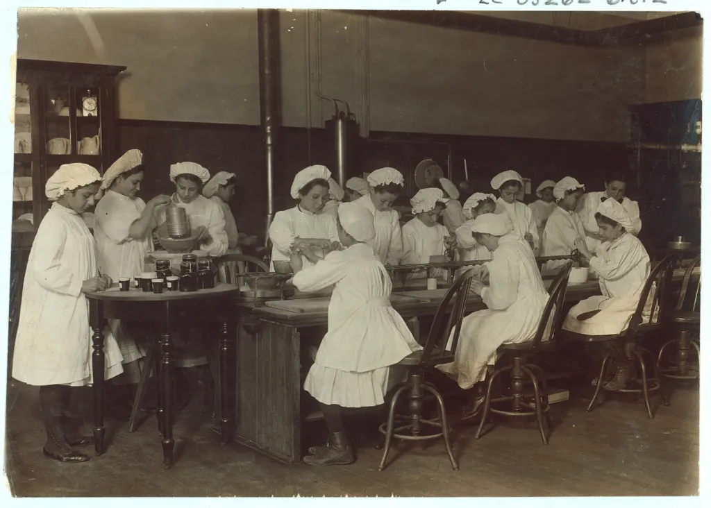 Early 20th-century black-and-white photo of girls in white caps and aprons working at long tables in a bread-making class, captioned Bread Making as Mother Makes It