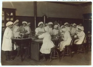 Early 20th-century black-and-white photo of girls in white caps and aprons working at long tables in a bread-making class, captioned Bread Making as Mother Makes It
