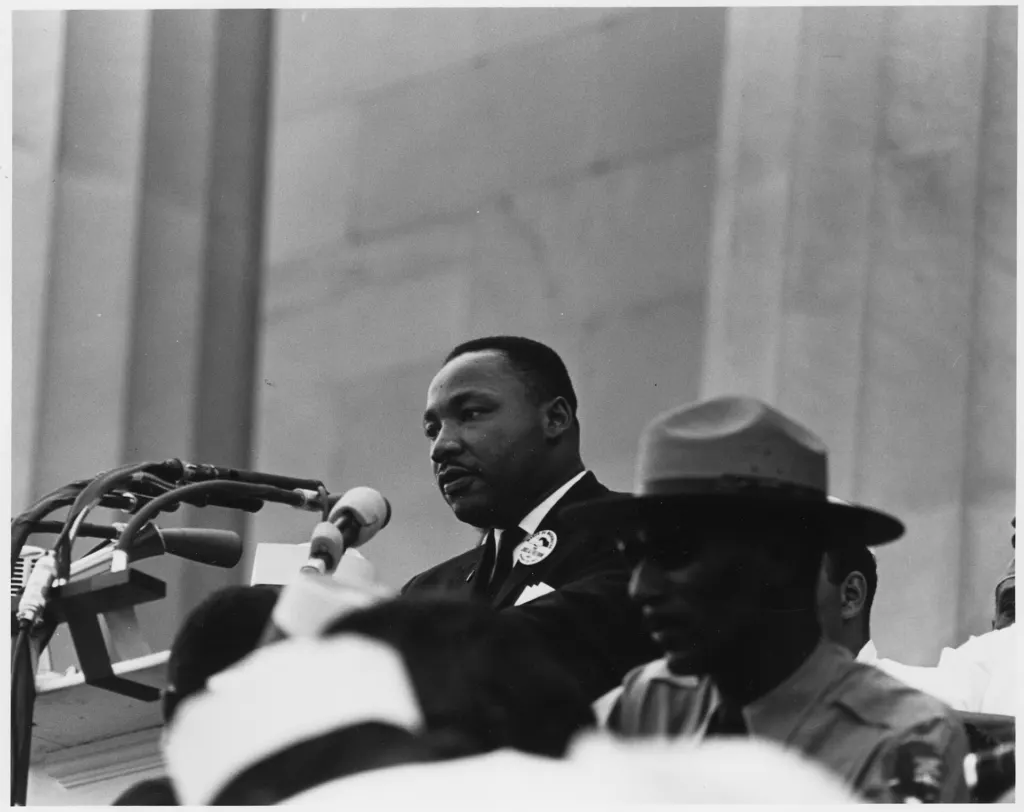 Martin Luther King Jr. speaking at a lectern crowded with microphones at the Lincoln Memorial during the March on Washington, August 28, 1963, with a man in a campaign hat in the foreground