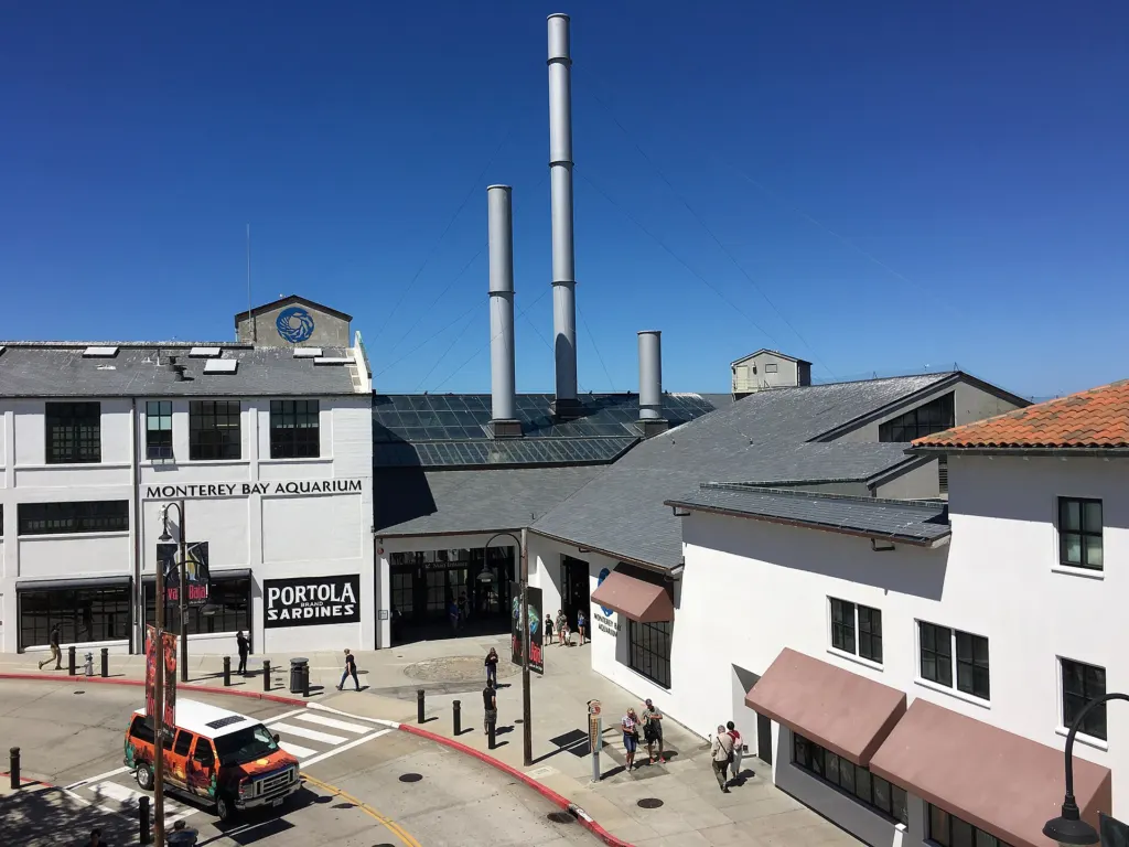 Main entrance of the Monterey Bay Aquarium on Cannery Row, with replica cannery smokestacks on the roof, seen from a bridge over the street