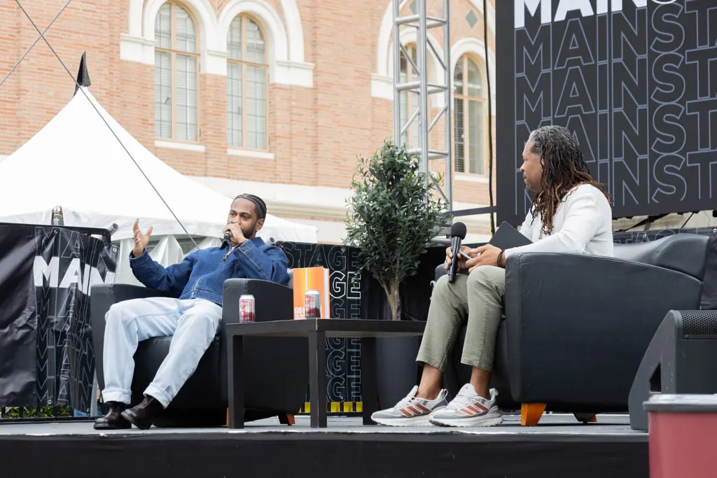Big Sean in a blue shirt and white jeans speaking into a microphone from an armchair on an outdoor stage at the Los Angeles Festival of Books in April 2025