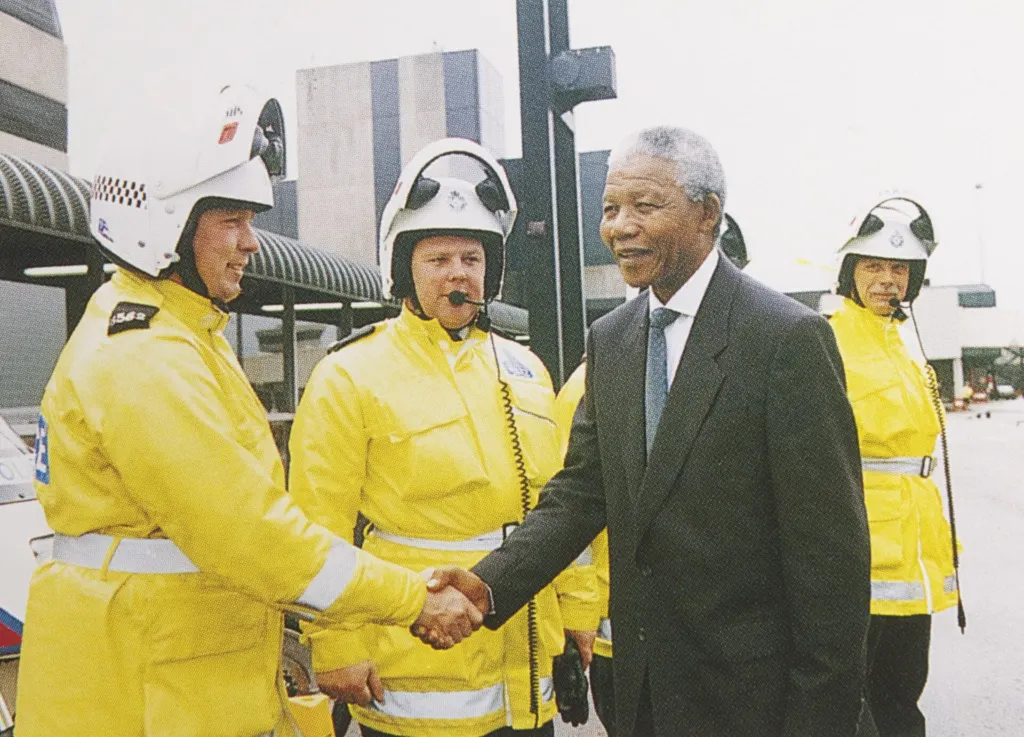 Nelson Mandela, in a dark suit and tie, smiles as he shakes hands with a police officer in a yellow high-visibility jacket and white helmet during his 1993 visit to the UK