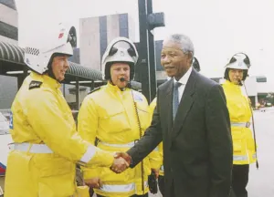 Nelson Mandela, in a dark suit and tie, smiles as he shakes hands with a police officer in a yellow high-visibility jacket and white helmet during his 1993 visit to the UK