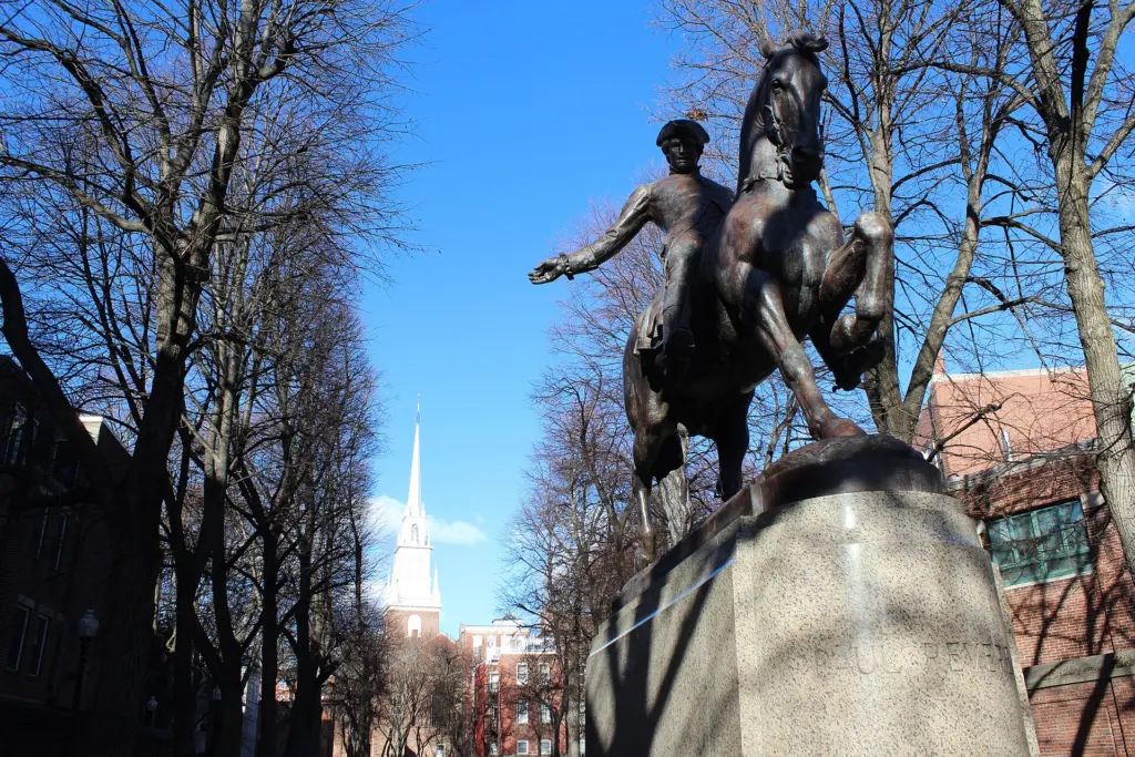 Bronze equestrian statue of Paul Revere on a stone pedestal in Boston's North End, with the white steeple of the Old North Church rising behind the trees