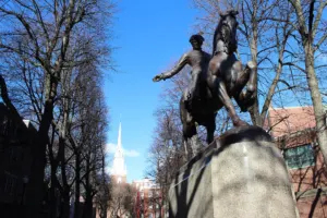 Bronze equestrian statue of Paul Revere on a stone pedestal in Boston's North End, with the white steeple of the Old North Church rising behind the trees