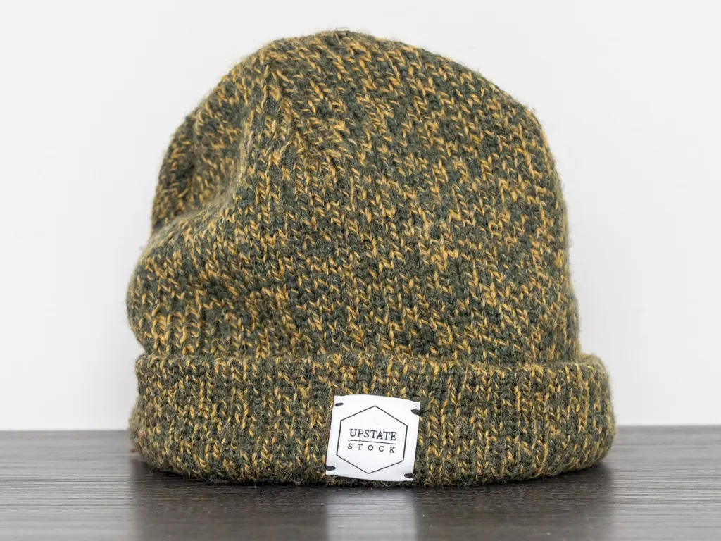 A green knit beanie with a small woven label on its cuff, sitting on a gray surface against a white background