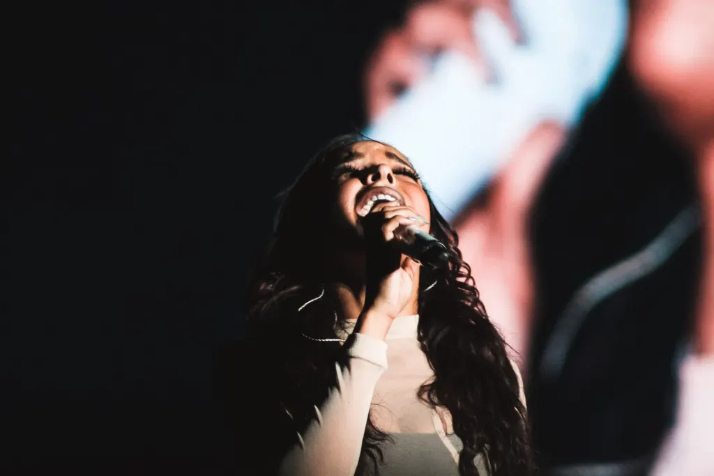 Tinashe singing into a handheld microphone with her head tilted back on a dark concert stage in 2015, a large video screen behind her