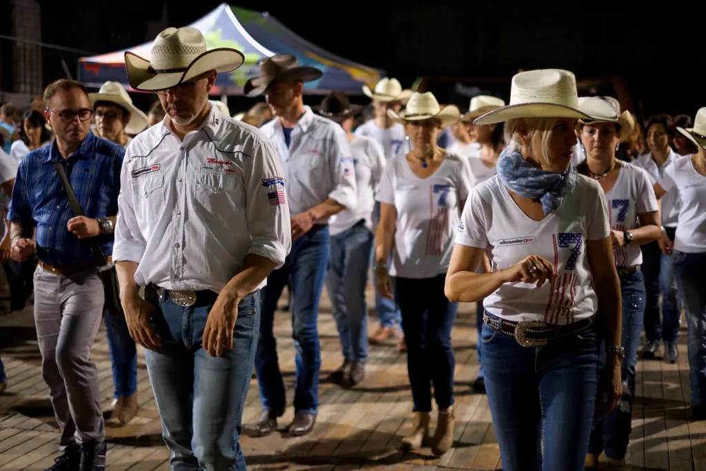 Rows of line dancers in cowboy hats, white shirts and jeans dancing at an outdoor evening event